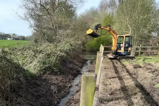 Pelleteuse jaune muni d’un broyeur forestier, ayant terminé le débroussaillage des herbes hautes et des ronces derrière une clôture.