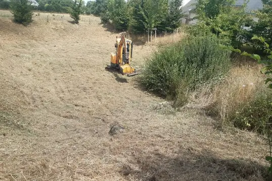 Une pelleteuse équipée d’un broyeur à végétation, sur un terrain envahi par des herbes hautes et des arbustes.