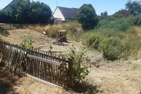 Une pelleteuse orange en train de débroussailler un terrain en pente, entouré d’herbes hautes et de quelques arbres, avec une clôture en bois au premier plan.