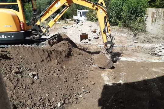 Travaux d’excavation à l’aide d’une pelleteuse dans un espace clos, avec gravats et tas de terre au premier plan.