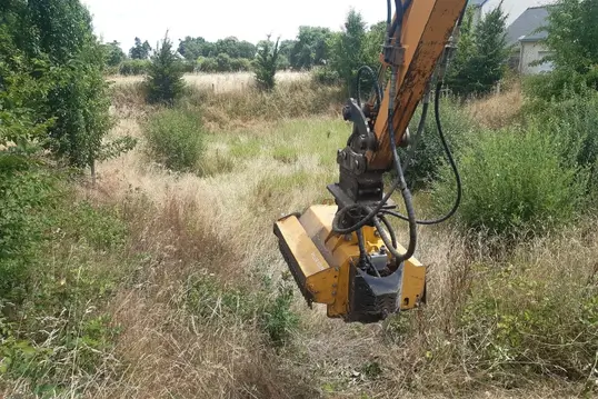 Bras hydraulique d’une pelleteuse équipé d’un broyeur à végétation, sur un terrain envahi par des herbes hautes et des arbustes.