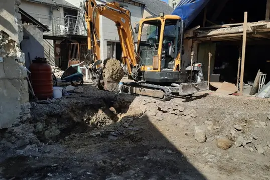 Bras hydraulique d’une mini-pelle déplaçant de la terre dans une cour en travaux, murs décrépis en arrière-plan.
