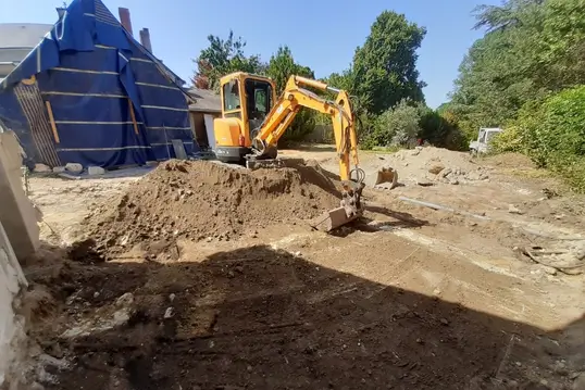 Mini-pelle compacte effectuant des travaux de terrassement devant un bâtiment partiellement couvert par une bâche bleue.