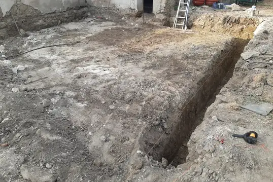 Tranchée creusée dans un terrain en terre pour des travaux de fondation, avec échelle posée à l’extrémité et outils de chantier au sol.