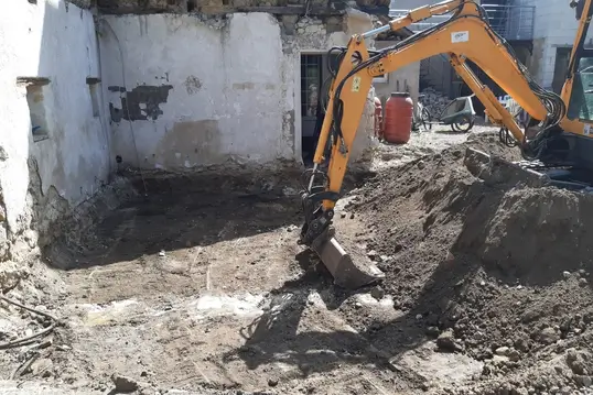 Pelleteuse en action creusant le sol d’un bâtiment en rénovation, avec murs anciens et matériel de chantier en arrière-plan.