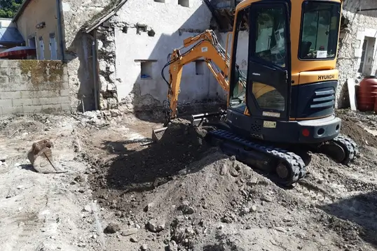 Mini-pelle excavatrice creusant à l’intérieur d’une structure partiellement démolie, sol dégagé et murs abîmés visibles.