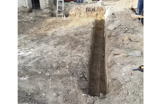 Vue sous un autre angle d’une tranchée de fondation creusée dans un terrain, avec échelle et matériel de chantier visibles en arrière-plan.